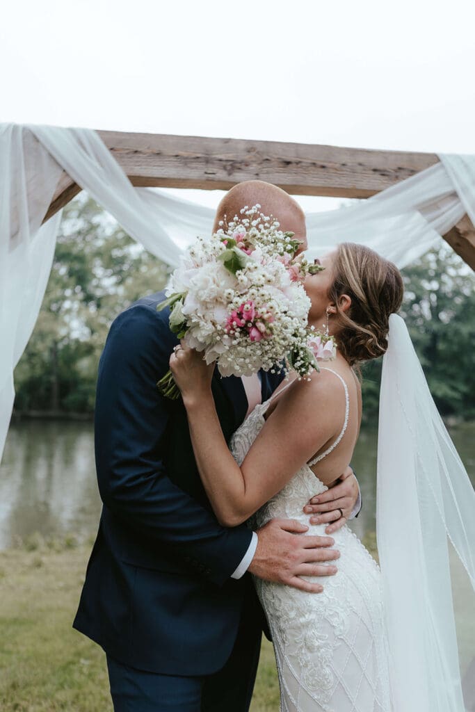 Bride and groom kiss under a wooden arch during their intimate wedding ceremony by a creek