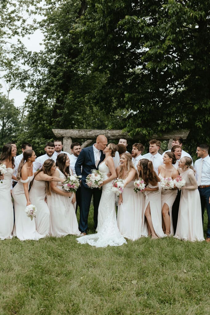 Bride and groom are surrounded by their entire wedding party, who is cheering while they kiss
