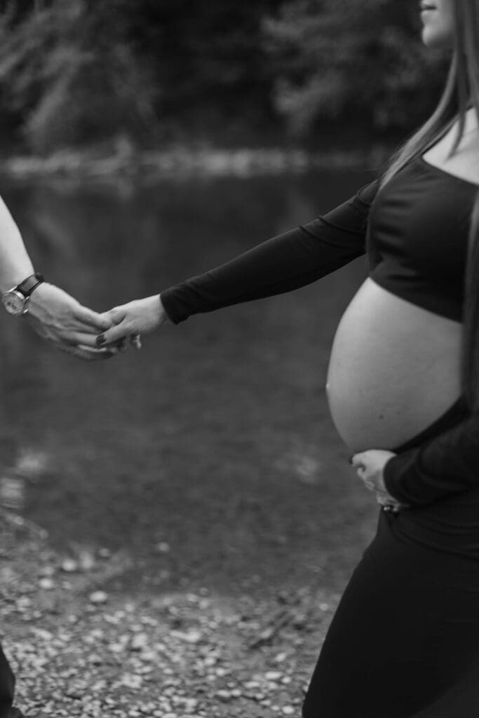 Woman holds belly and holds the hand of her husband as they walk on the banks of a creek