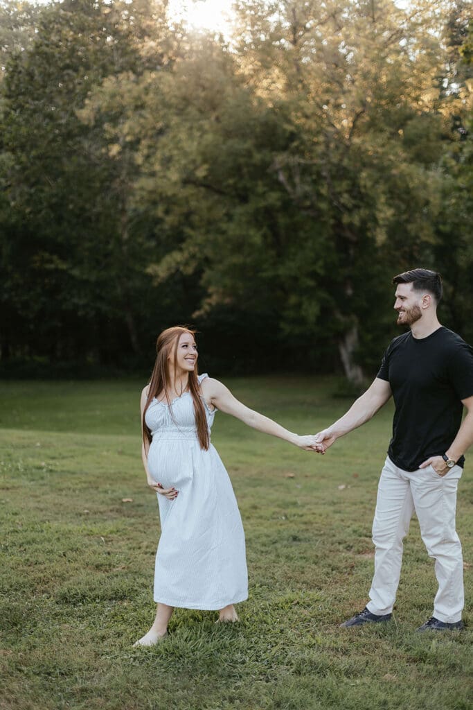 Woman holds her belly and holds the hand of her husband while walking through a field