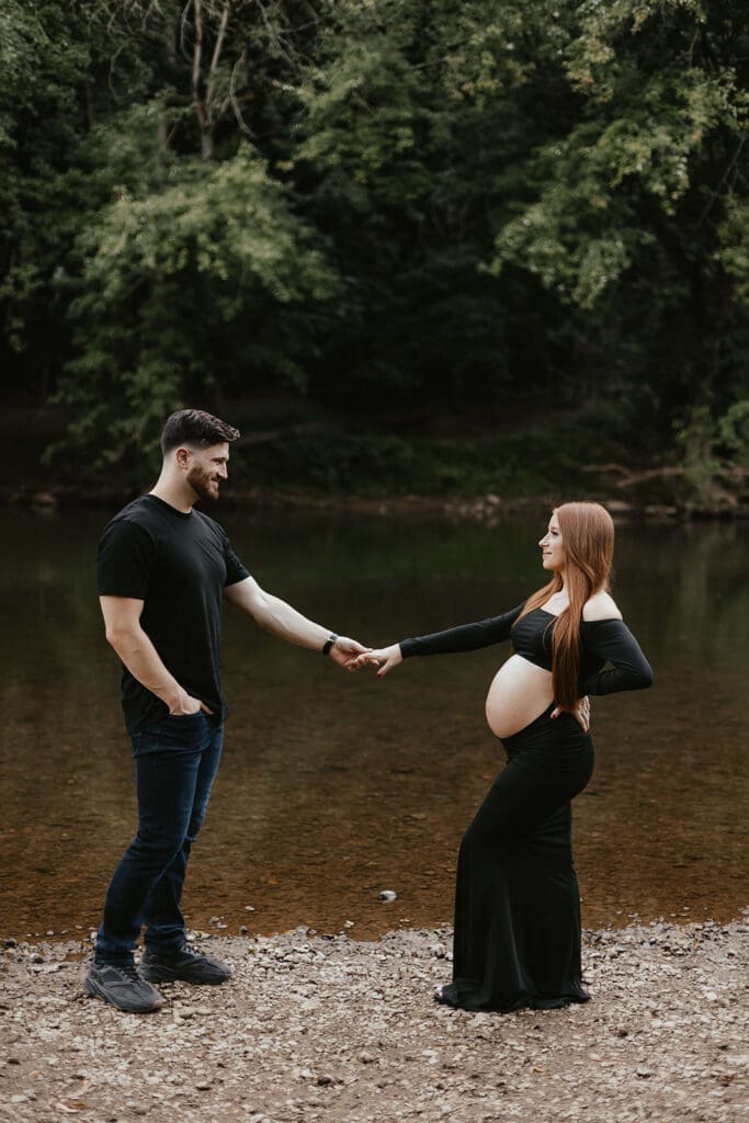 Man and woman holding hands during their outdoor maternity photos by a creek