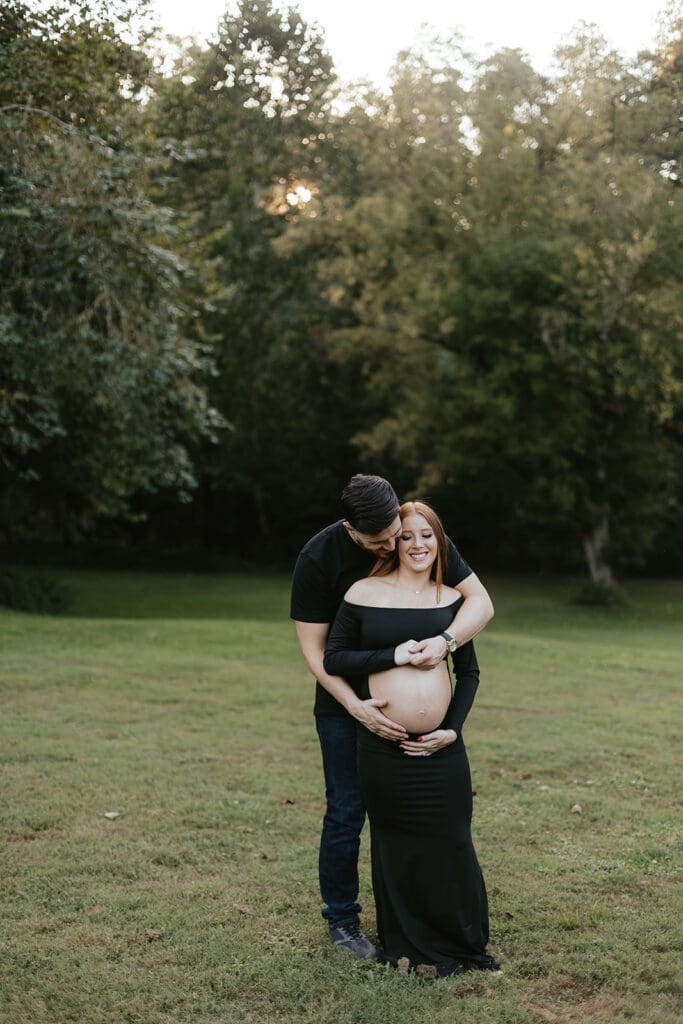 Man holds pregnant wife's stomach in a field while the wife smiles