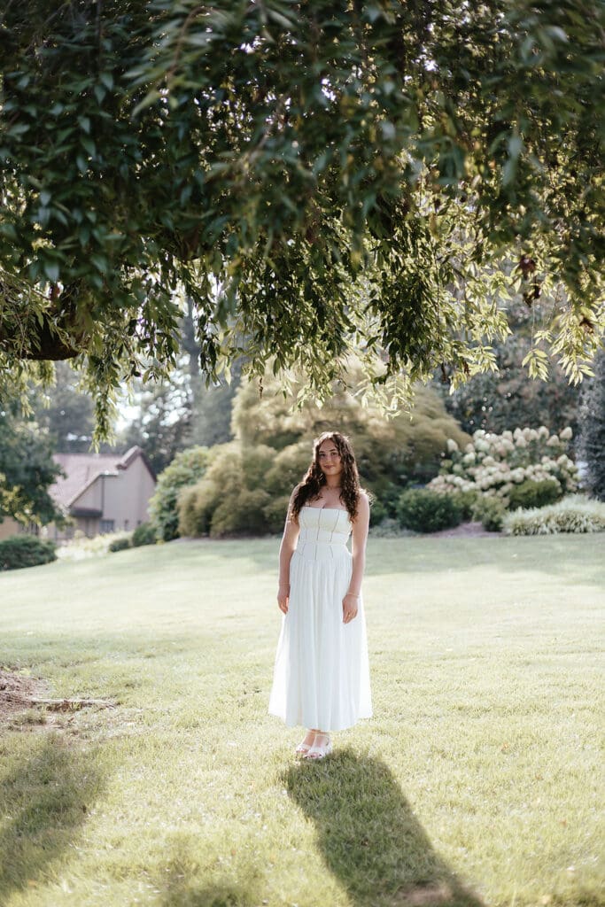 Senior in long, flowing white dress stands under a tree in a backyard