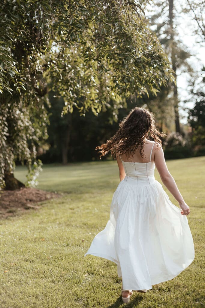 Senior in long, flowing white dress walks through an open field surrounded by trees