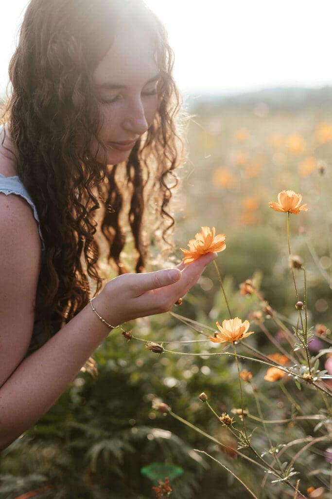 Senior with long curly hair touches an orange flower in a field of wildflowers