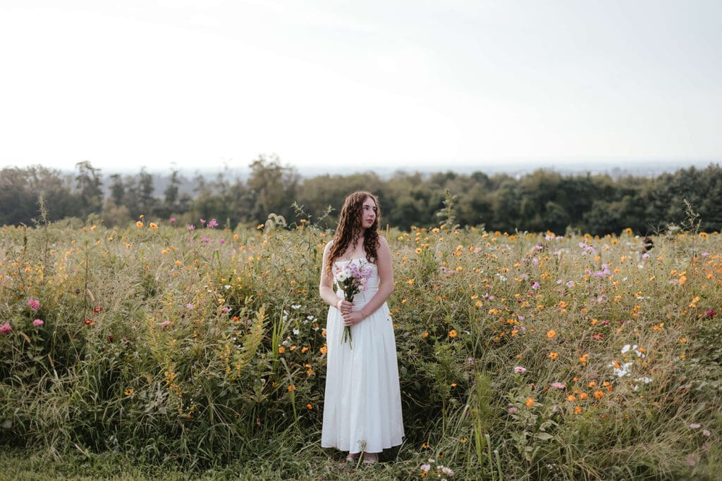 Senior in a long white dress and bouquet stands in a field at Wildflower Lookout in Lancaster