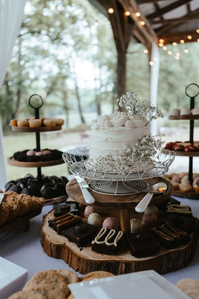 White wedding cake on wooden stand surrounded by brownies and other desserts