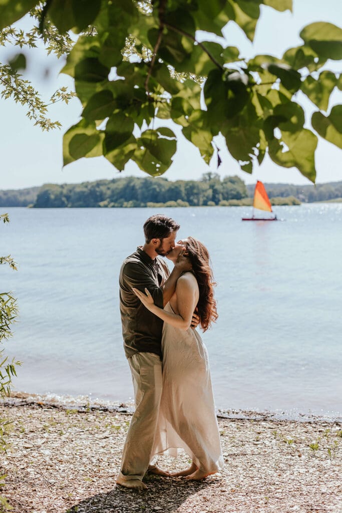 Romantic image of a man and woman kissing in front of a lake with a colorful sailboat in the background