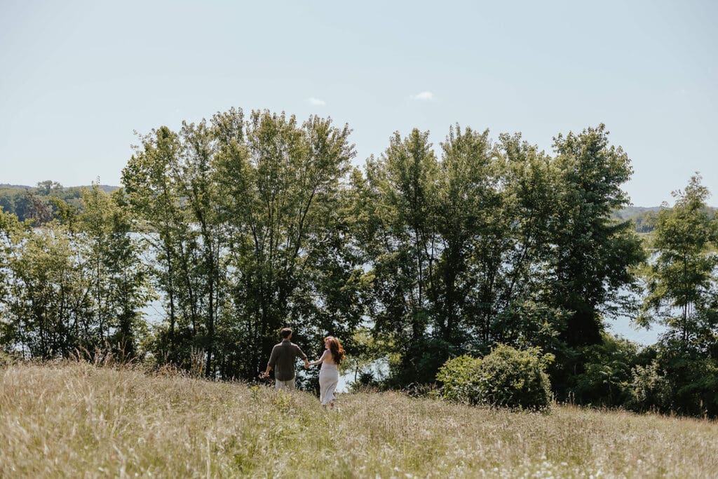 Summer engagement photo of a couple running downhill through a field towards a lake