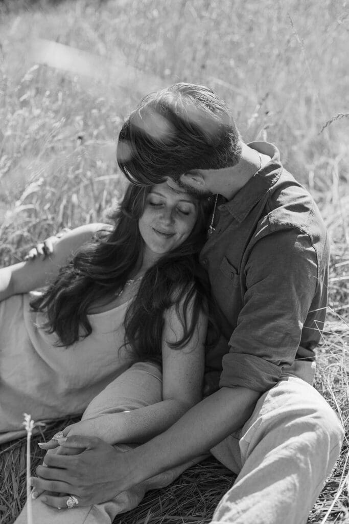 Man kisses his fiancée's forehead as they lie on the grass during their summer engagement photos