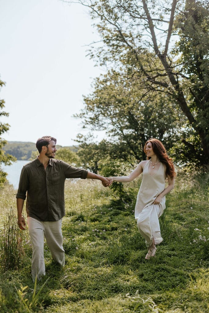 Engaged couple walks holding hands through a field by a lake