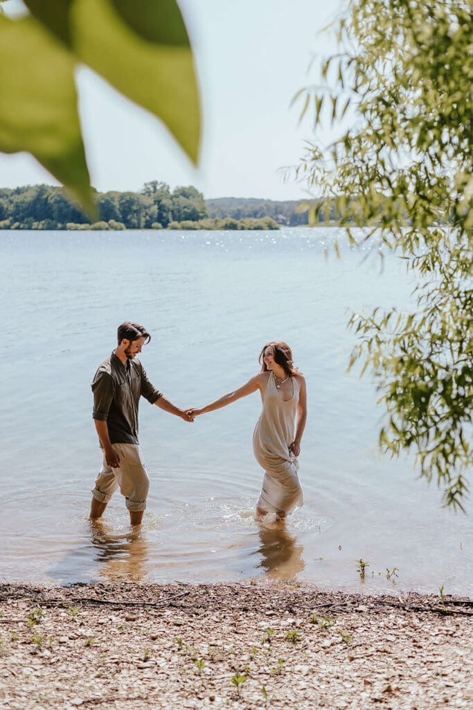 Summer candid engagement photo of a couple holding hands while walking barefoot through a lake