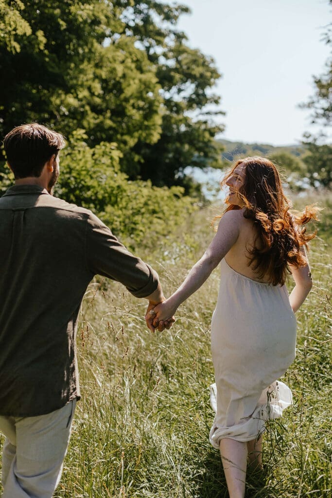 Engaged couple runs holding hands through a field by a lake