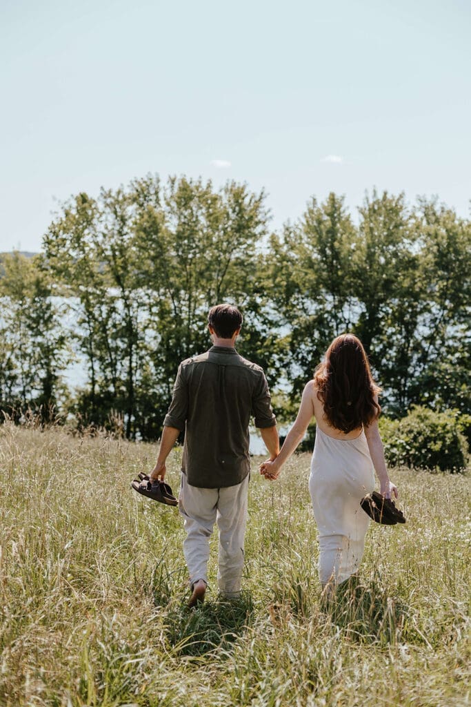 Summer engagement photo of an engaged couple walking holding hands through a field by a lake