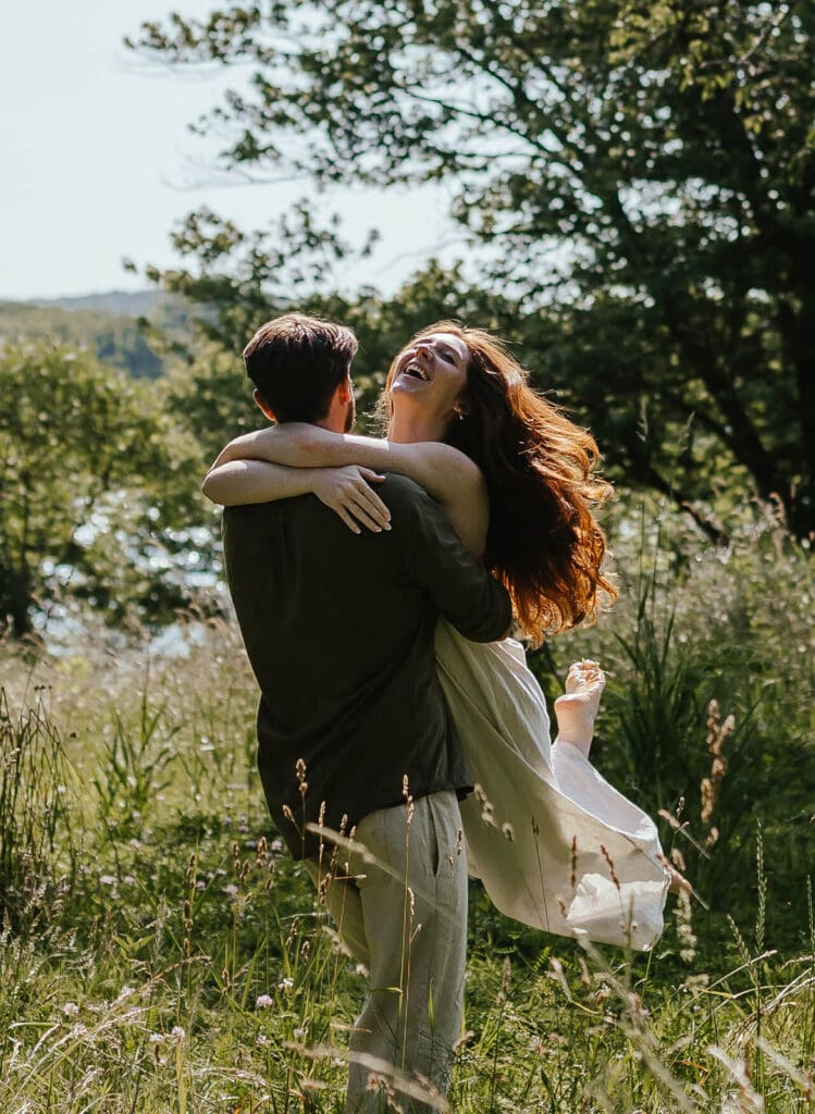Candid photo of woman laughing as her fiancé swings her in an open field in the summer
