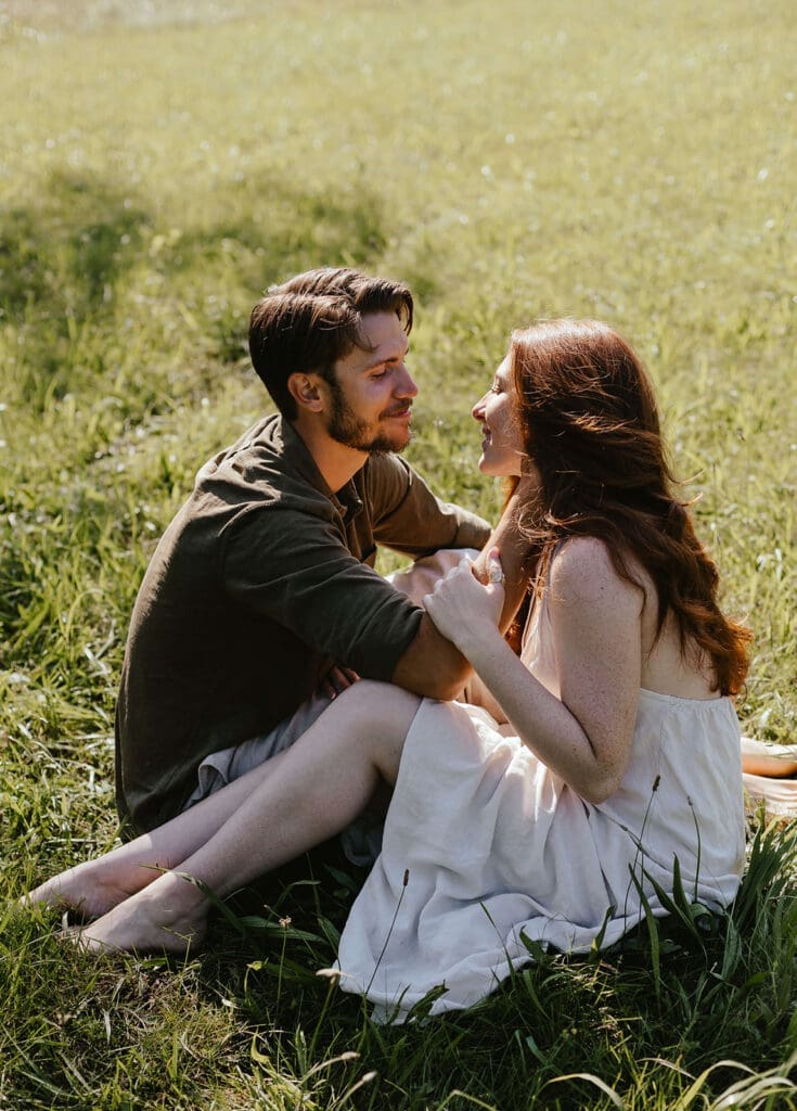 Summer engagement photos of couple gazing lovingly into each other's eyes while sitting in a field
